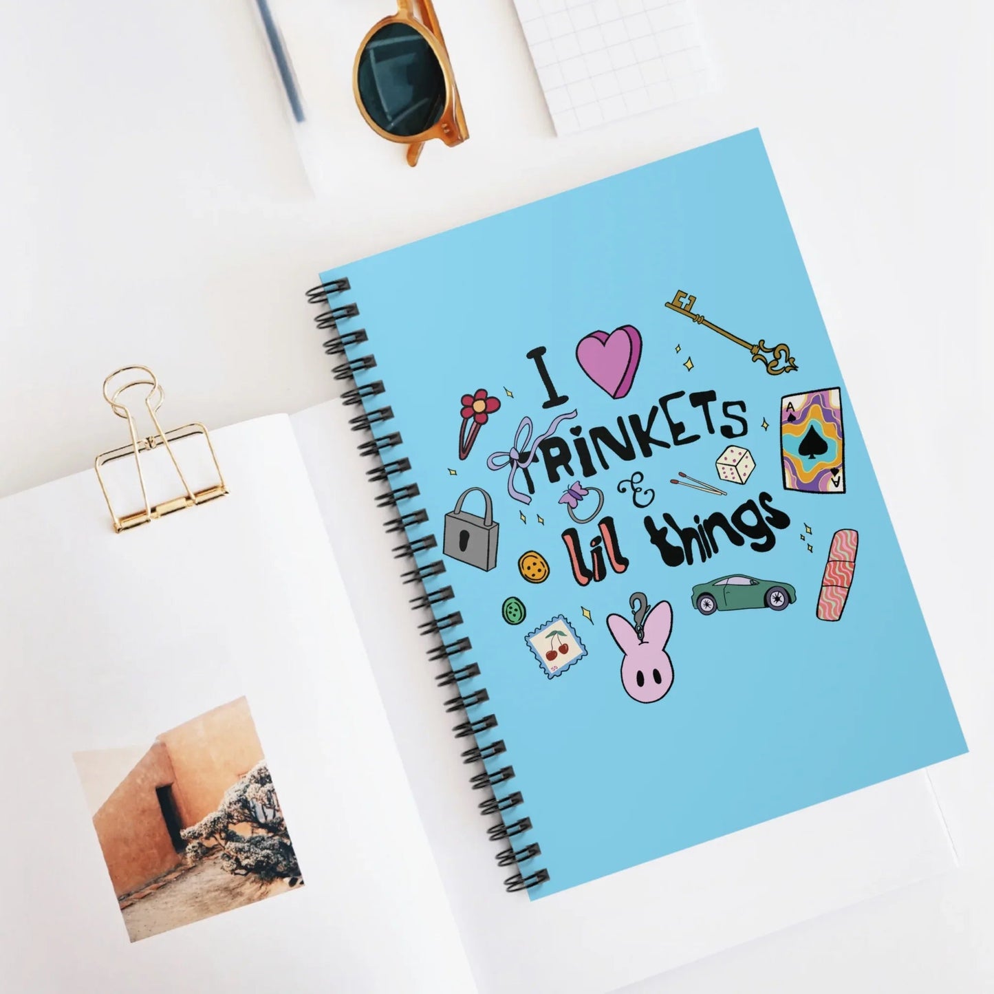 A blue notebook with doodles and the phrase "I ♥ trinkets & lil things" on the cover. Surrounding items include a key, dice, and a bunny illustration, conveying a playful vibe.