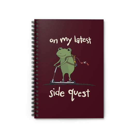 A whimsical cartoon frog rides a scooter on a spiral notebook cover, with the text "on my latest side quest" in playful white font against a durable maroon notebook cover.