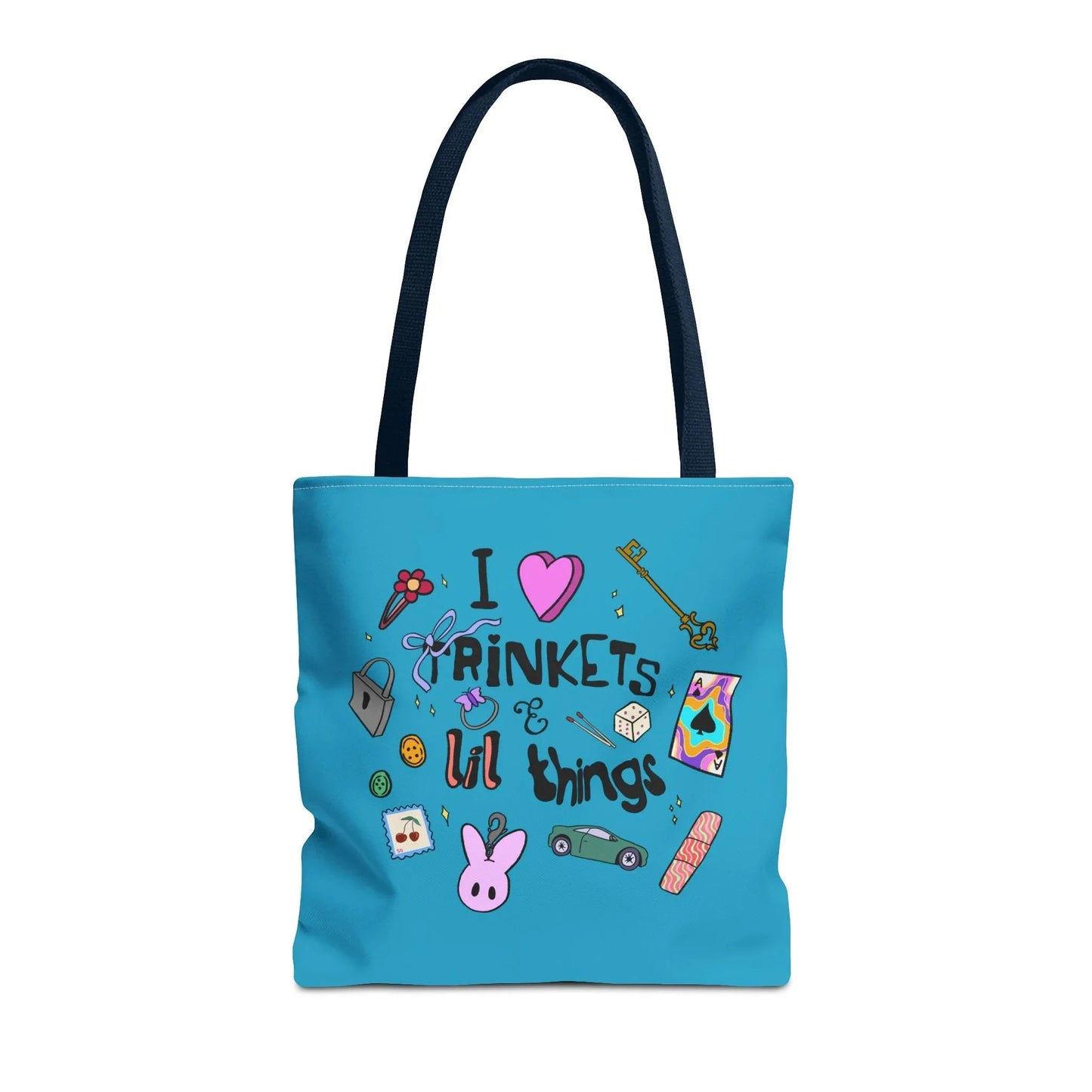Blue tote bag with navy handles, featuring playful doodles like a key, ribbon, dice, and cartoon bunny. Text reads "I love trinkets & lil things." Fun and whimsical.