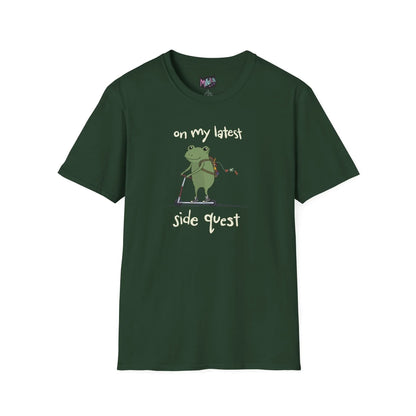 Green t-shirt with a cute green frog on a scooter, wearing a backpack that is slightly unzipped with small items hanging out. Text reads, "on my latest side quest." Whimsical and playful design.