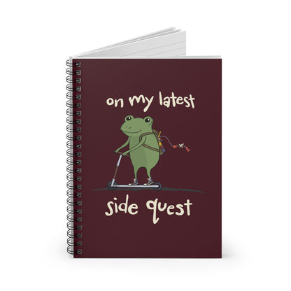 A whimsical cartoon frog rides a scooter on a spiral notebook cover, with the text "on my latest side quest" in playful white font against a durable maroon notebook cover.
