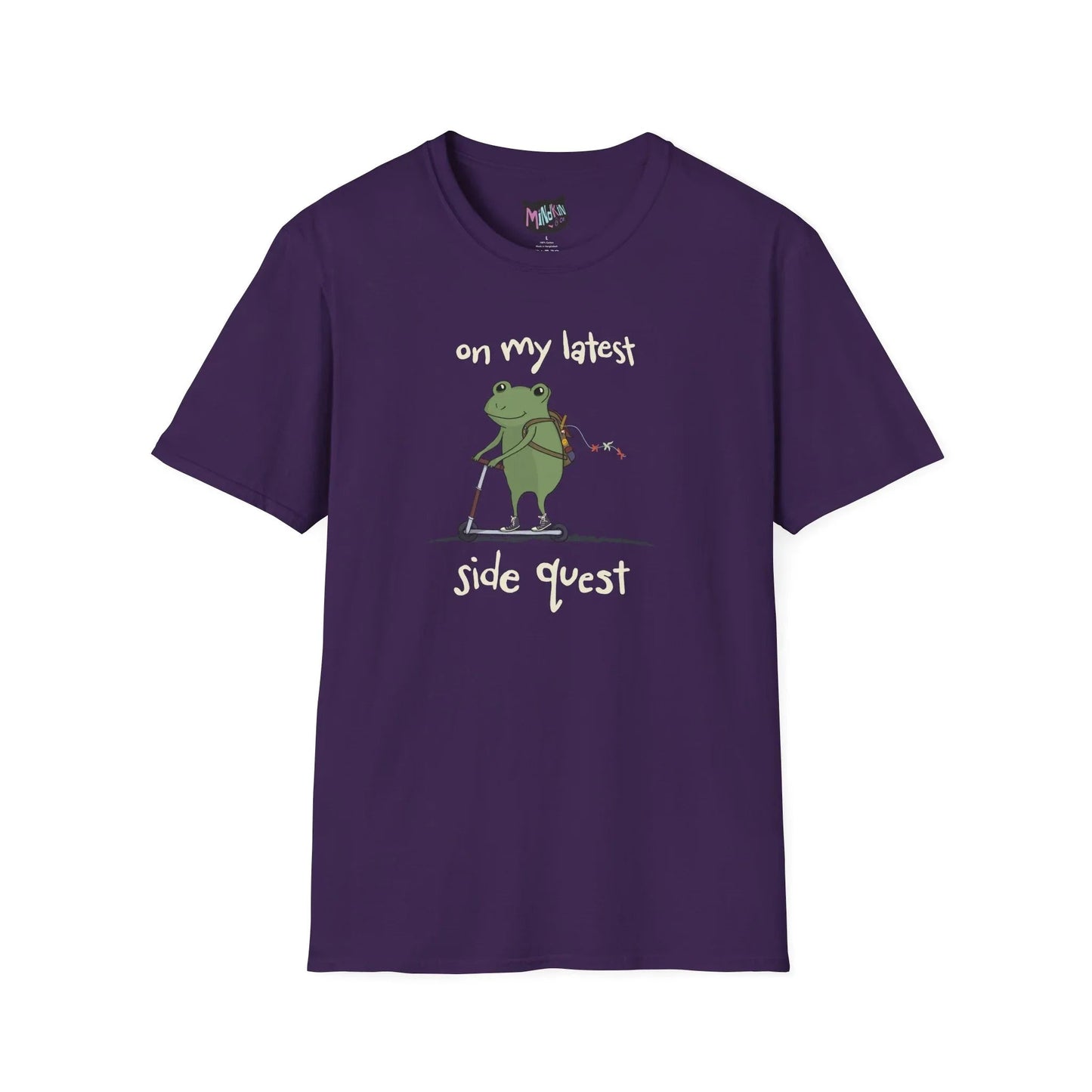 Purple t-shirt with a cute green frog on a scooter, wearing a backpack that is slightly unzipped with small items hanging out. Text reads, "on my latest side quest." Whimsical and playful design.