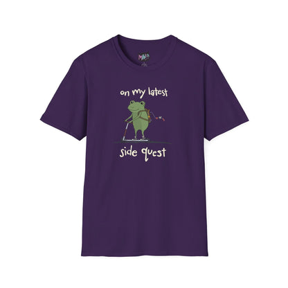 Purple t-shirt with a cute green frog on a scooter, wearing a backpack that is slightly unzipped with small items hanging out. Text reads, "on my latest side quest." Whimsical and playful design.