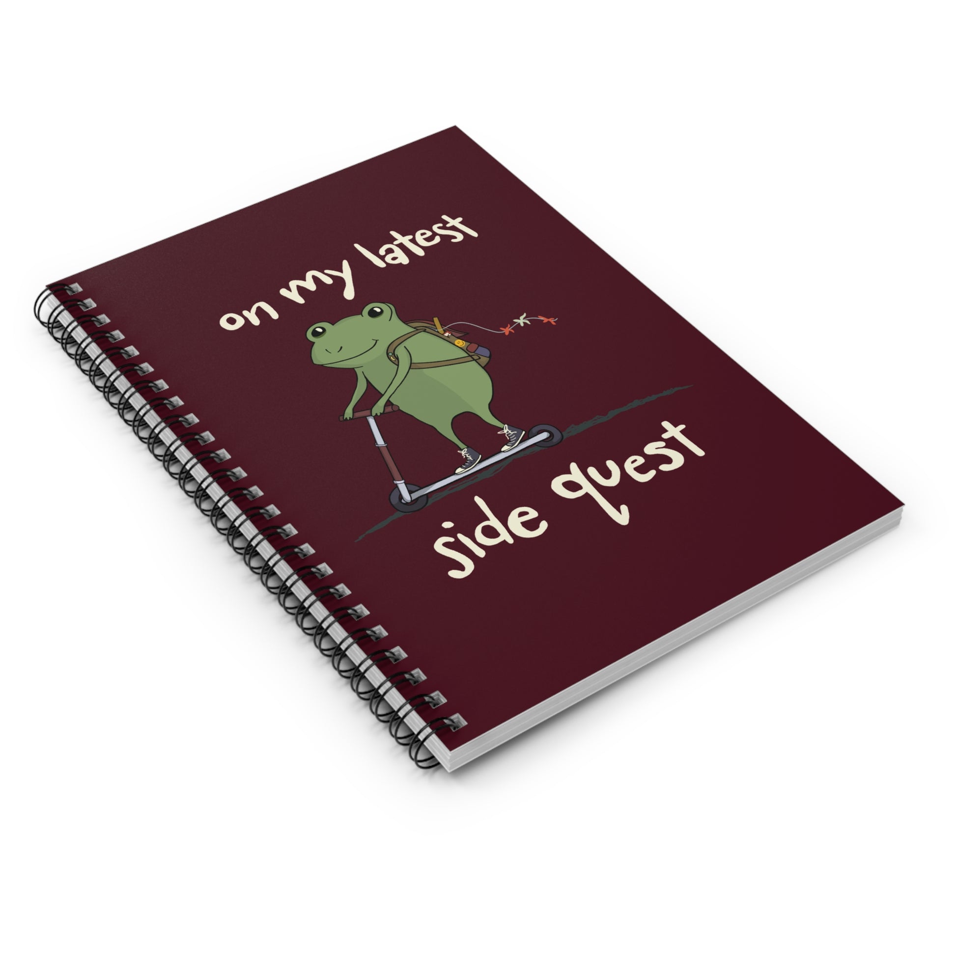 Spiral notebook with a maroon cover featuring a cheerful cartoon frog on a scooter. Metal spiral binding lays flat and allows easy writing.