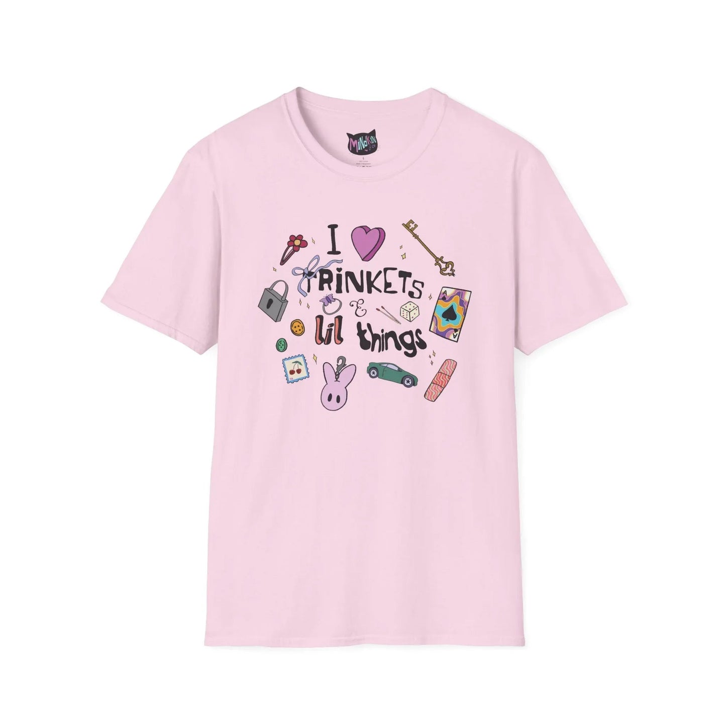 Pink T-shirt featuring doodles of ornate keys, dice, a toy car, a flower hairclip, a postage stamp, a butterfly ring, and a pink bunny keychain. Text reads "I love trinkets and lil things" with playful, colorful illustrations.