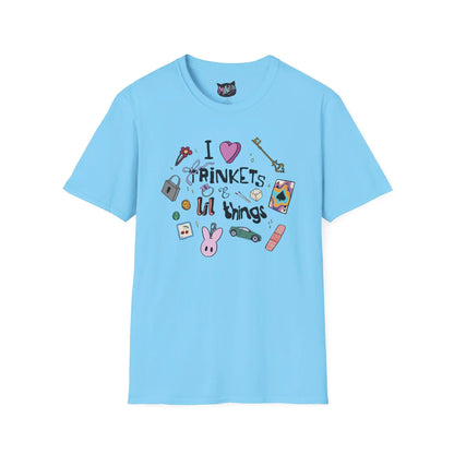Blue T-shirt featuring doodles of ornate keys, dice, a toy car, a flower hairclip, a postage stamp, a butterfly ring, and a pink bunny keychain. Text reads "I love trinkets and lil things" with playful, colorful illustrations.
