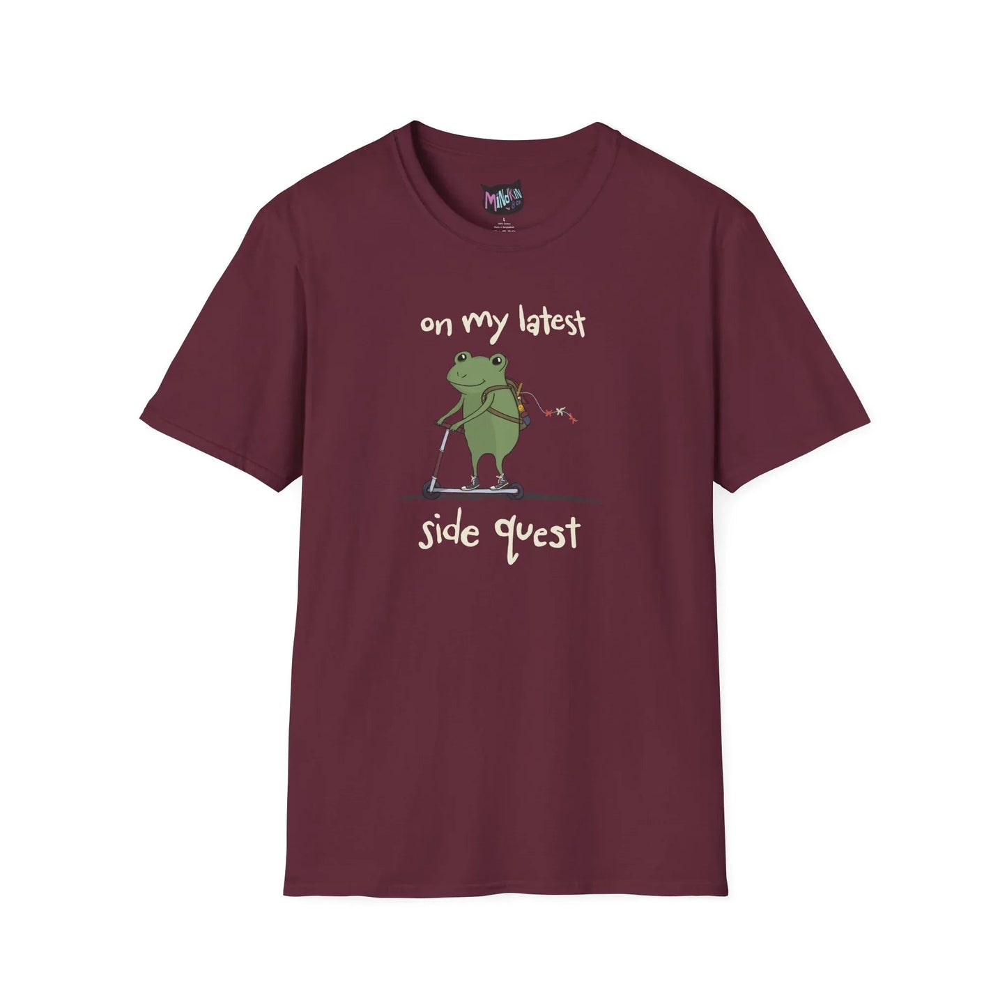 Maroon t-shirt with a cute green frog on a scooter, wearing a backpack that is slightly unzipped with small items hanging out. Text reads, "on my latest side quest." Whimsical and playful design.