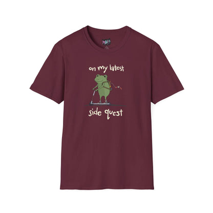 Maroon t-shirt with a cute green frog on a scooter, wearing a backpack that is slightly unzipped with small items hanging out. Text reads, "on my latest side quest." Whimsical and playful design.