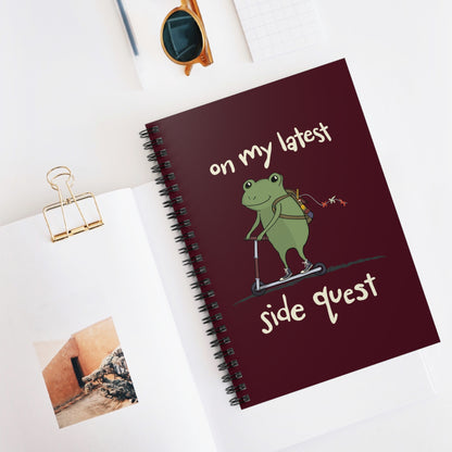 A spiral-bound notebook with a maroon cover featuring a cartoon frog riding a scooter. The text reads "on my latest side quest." 