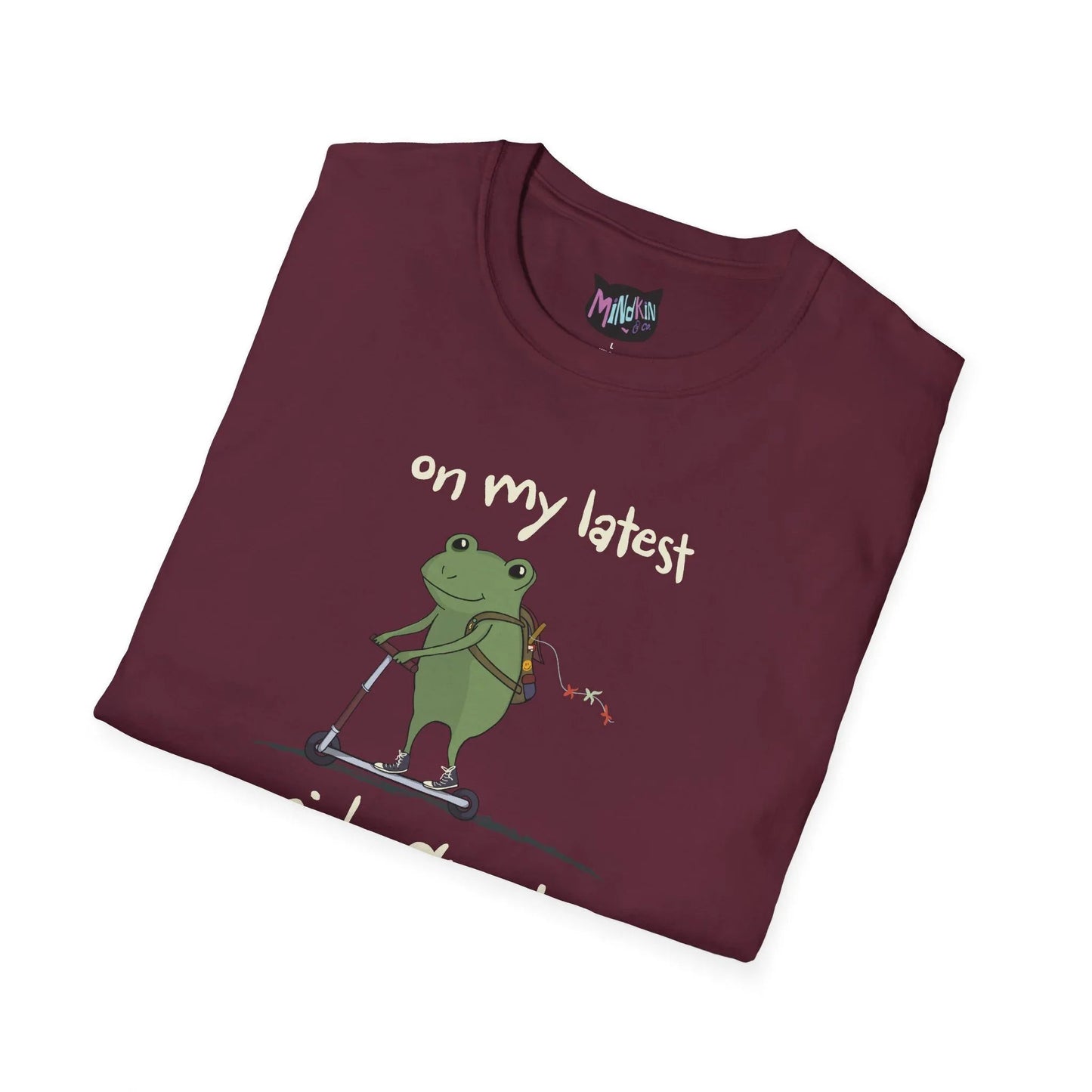 A folded maroon t-shirt featuring a cartoon frog riding a scooter with a backpack. The text reads "on my latest." The tone is playful and whimsical.