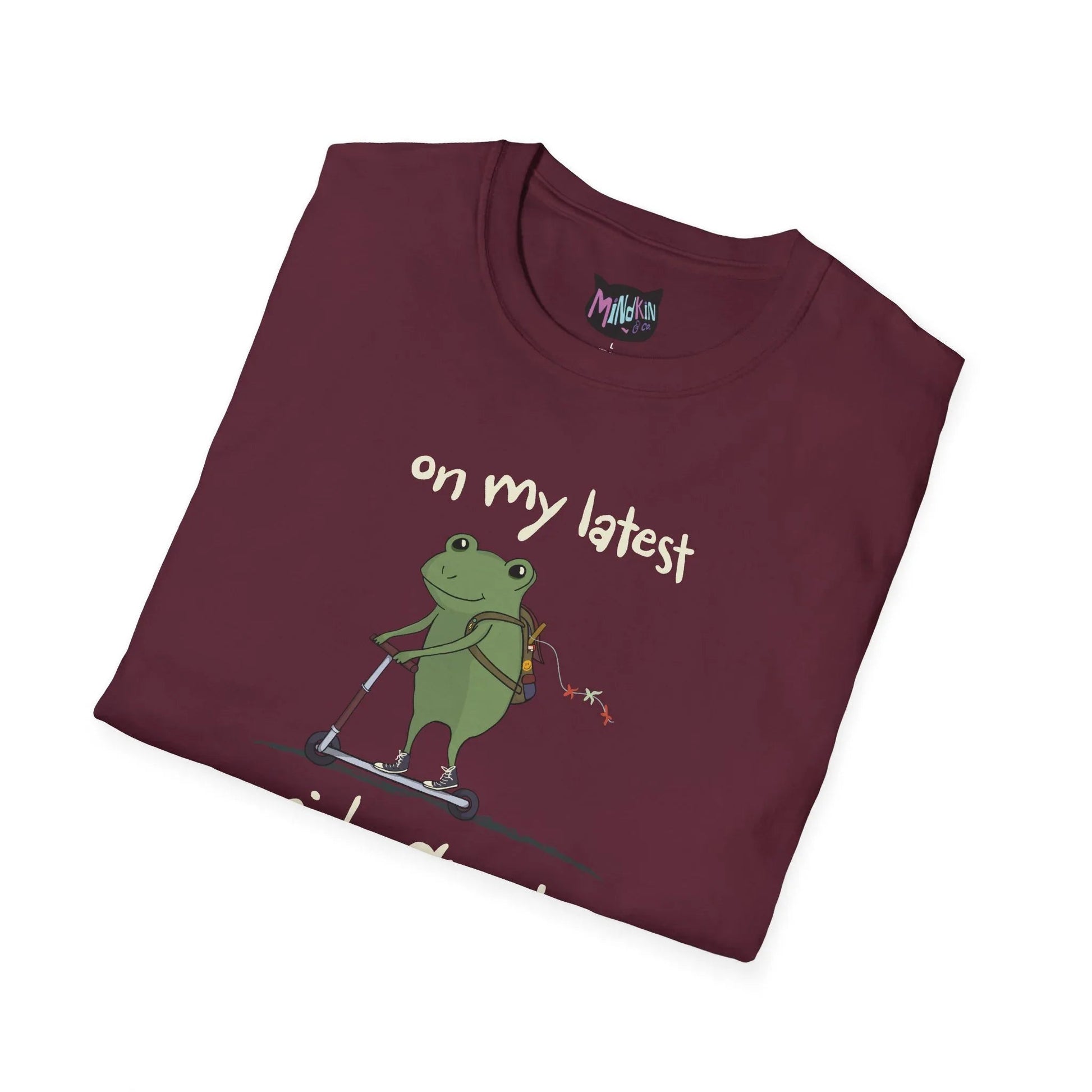 A folded maroon t-shirt featuring a cartoon frog riding a scooter with a backpack. The text reads "on my latest." The tone is playful and whimsical.