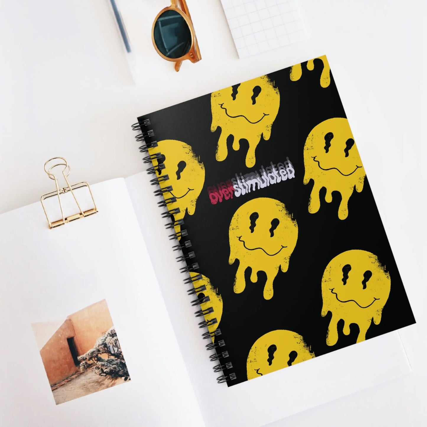 Spiral notebook with a black cover featuring multiple dripping yellow smiley faces and the word "Overstimulated."