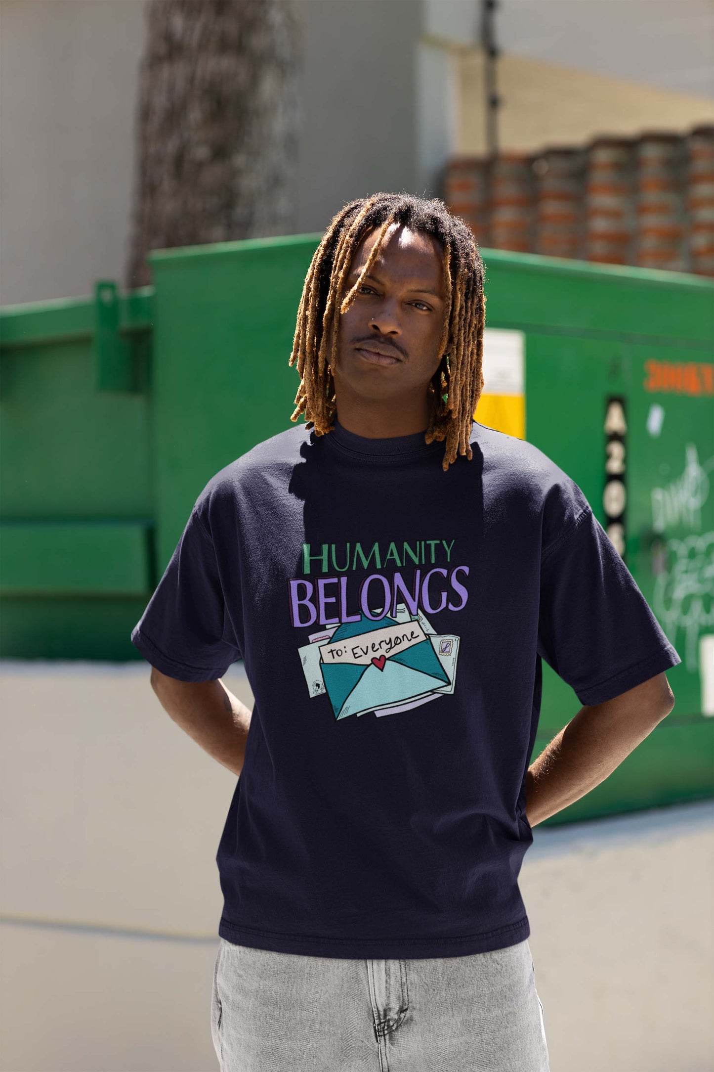 A person stands in front of a green dumpster, wearing a navy shirt with the message "Humanity Belongs to Everyone." They appear thoughtful and confident.