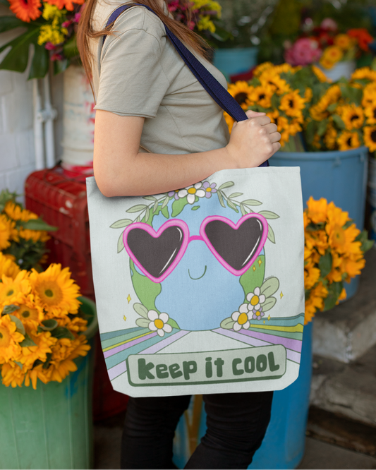 Woman in a flower shop holds a sturdy Keep It Cool Earth tote bag, with a cute Earth design wearing pink heart sunglasses. Text reads "keep it cool." at the bottom underneath the planet. 