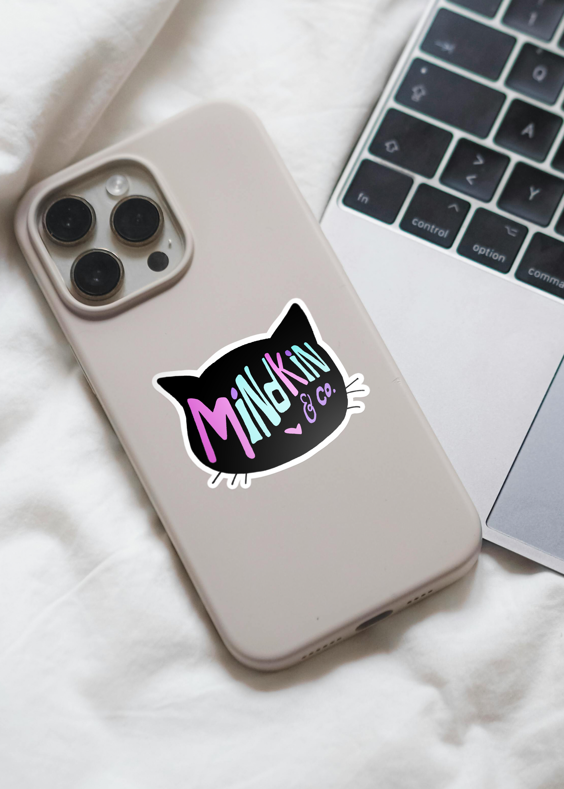 Smartphone with a white case featuring a Mindkin and Co. sticker on the back, next to an open laptop.