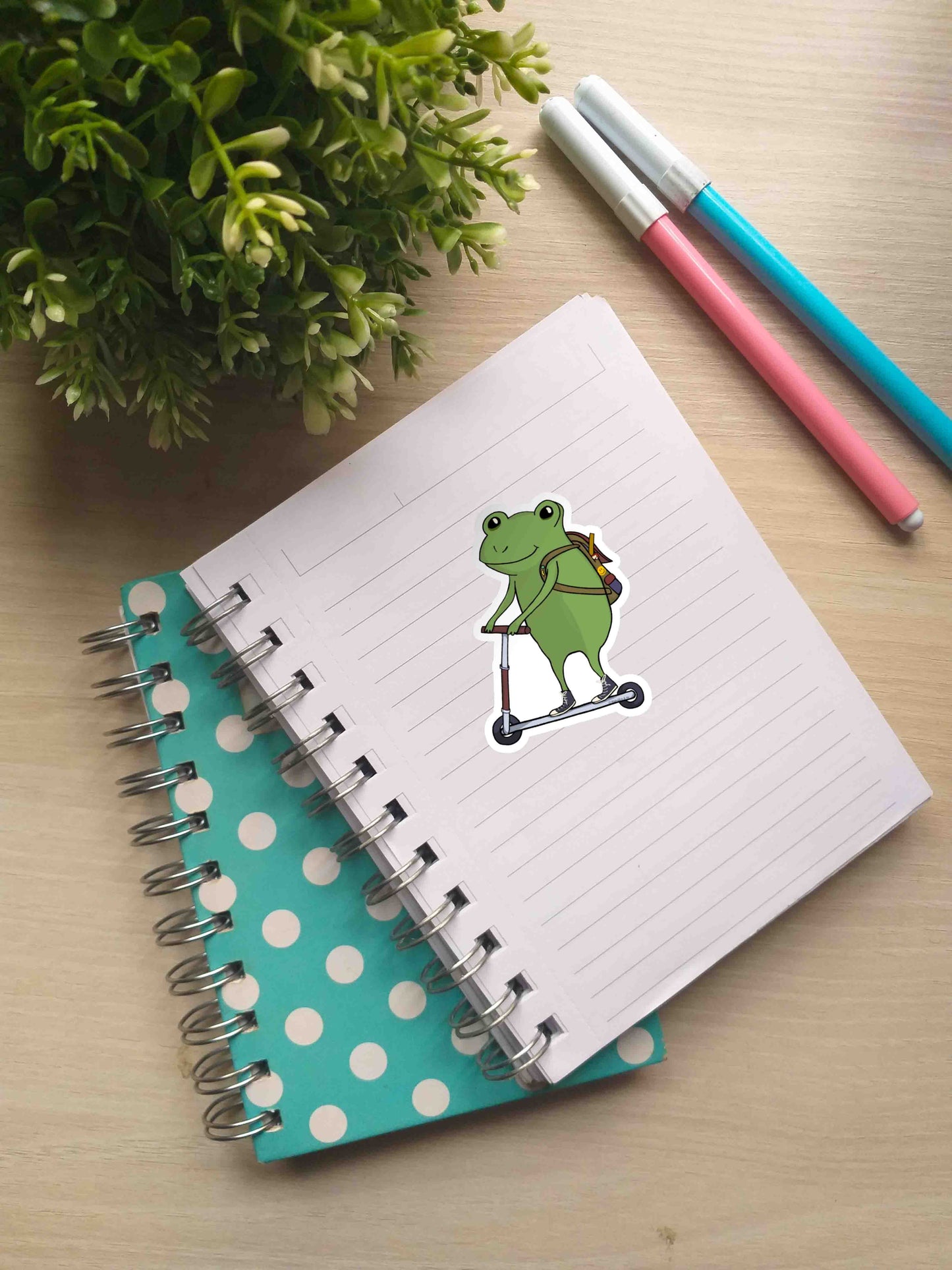 A notebook with Side Quest Frog sticker on a page. Nearby, there are two markers and a small potted plant.