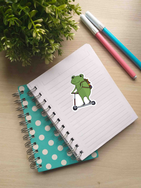 A notebook with Side Quest Frog sticker on a page. Nearby, there are two markers and a small potted plant.