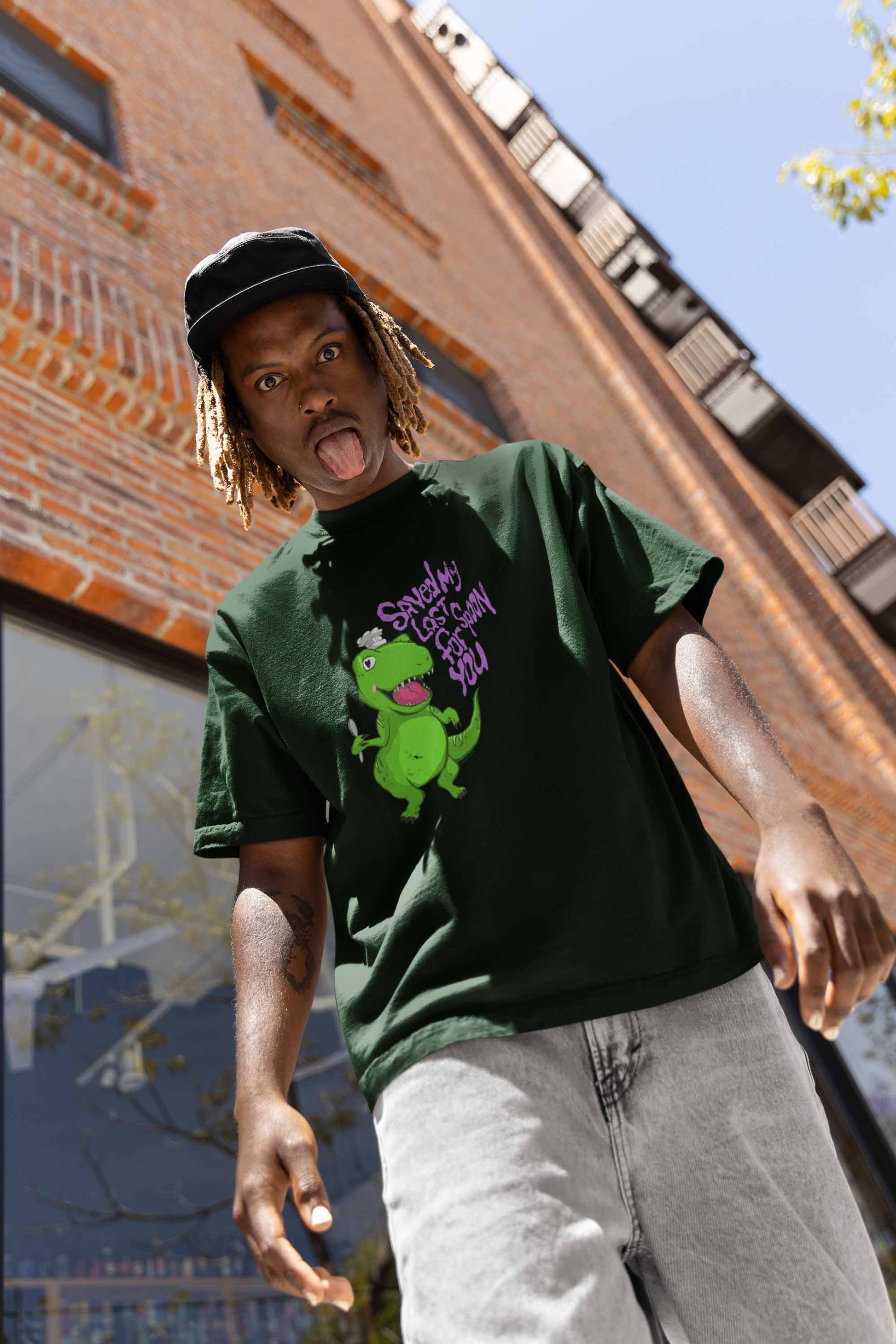 Young person with a playful expression sticks out their tongue, wearing a dark green Spoonie Cute Dino T-shirt. Red brick building in background.
