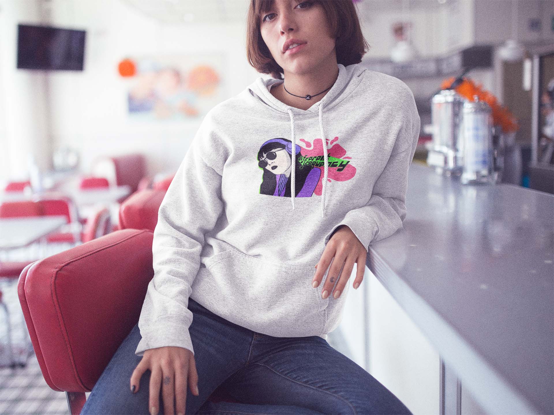 Young person in a casual diner setting, wearing a light gray Different Frequency. They sit on a red barstool, conveying a relaxed vibe.
