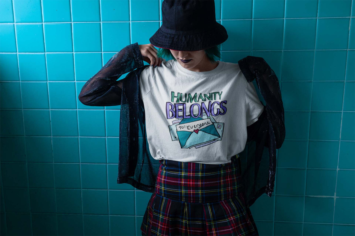 A person in a plaid skirt, black hat, and sheer jacket reveals a shirt with "Humanity Belongs to Everyone" text. Blue-tiled background; tone is bold and expressive.