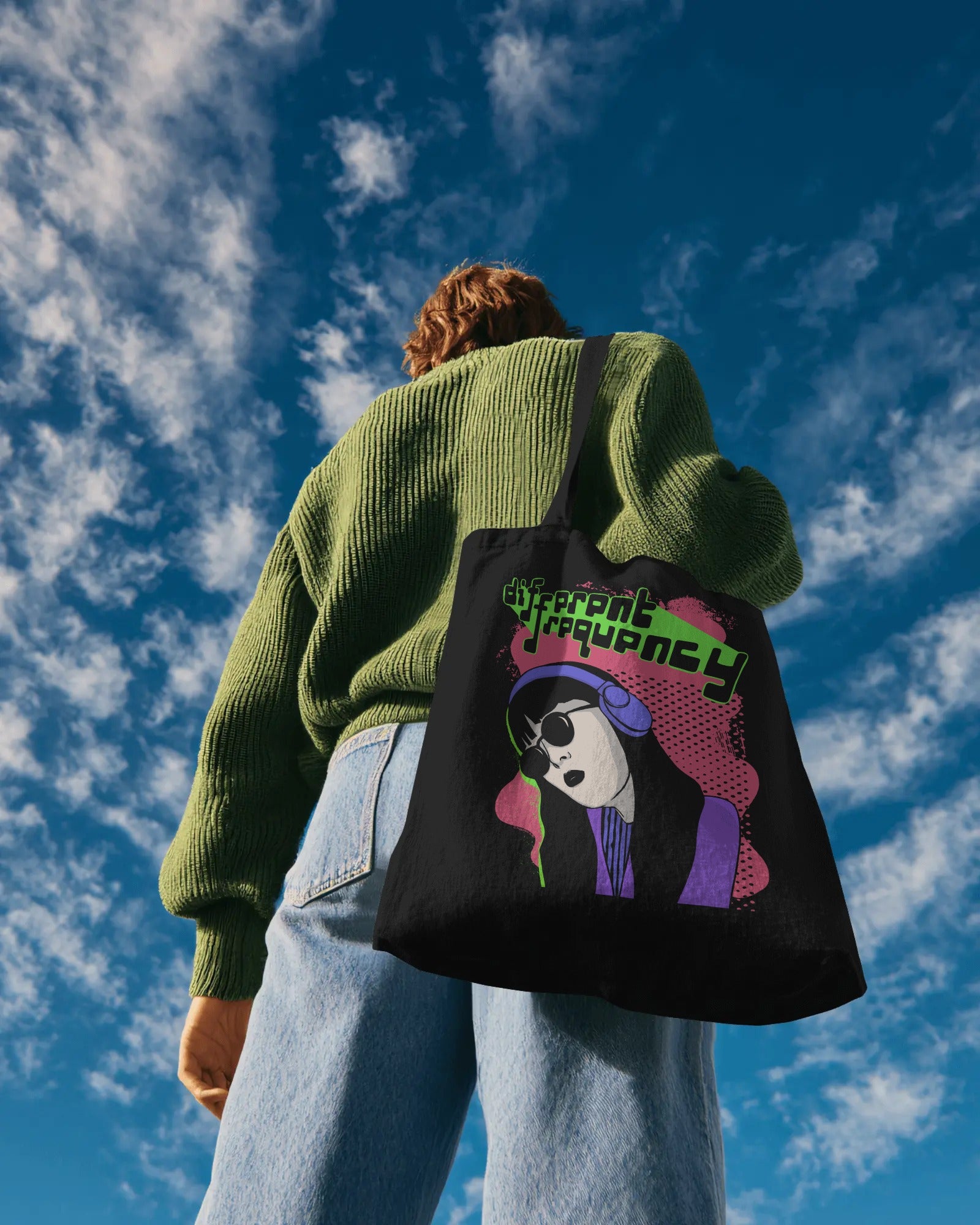 Person in a green sweater and jeans stands against a blue sky, holding a black tote bag with a vibrant illustration and the text "Different Frequency."